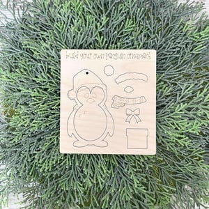 May include: A wooden craft kit for making a penguin ornament. The kit includes a penguin body, hat, scarf, bow, and gift box. The text "Build your own penguin ornament!" is printed on the wood.