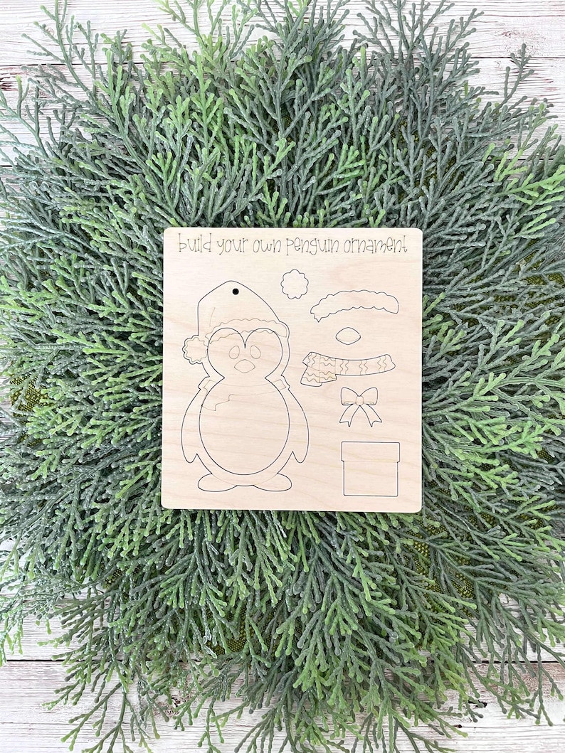 May include: A wooden craft kit for making a penguin ornament. The kit includes a penguin body, hat, scarf, bow, and gift box. The text "Build your own penguin ornament!" is printed on the wood.