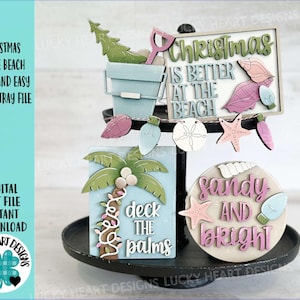 May include: A tiered tray decoration with a beach Christmas theme. It features a sign that says "Christmas is better at the beach", a palm tree, seashells, and the phrase "sandy and bright". The color palette includes blue, green, pink, and white.