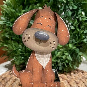 May include: A wooden dog figurine with a brown body, large ears, and a gray nose. The dog has white speckles and a white chest. It is sitting on a small wooden base. The background features green foliage.