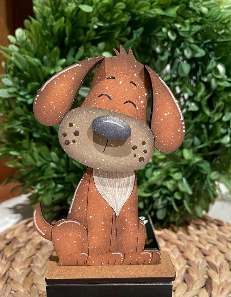 May include: A wooden dog figurine with a brown body, large ears, and a gray nose. The dog has white speckles and a white chest. It is sitting on a small wooden base. The background features green foliage.