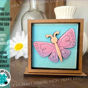 May include: A small wooden box with a light blue background and a pink and purple butterfly cutout. The box is standing on a wooden base. The text "LUCKY HEART DESIGNS" is printed on the base.