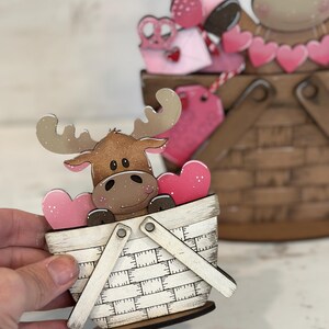 May include: A wooden moose figurine holding pink hearts in a white and brown basket with a white handle. The basket is decorated with a checkered pattern.