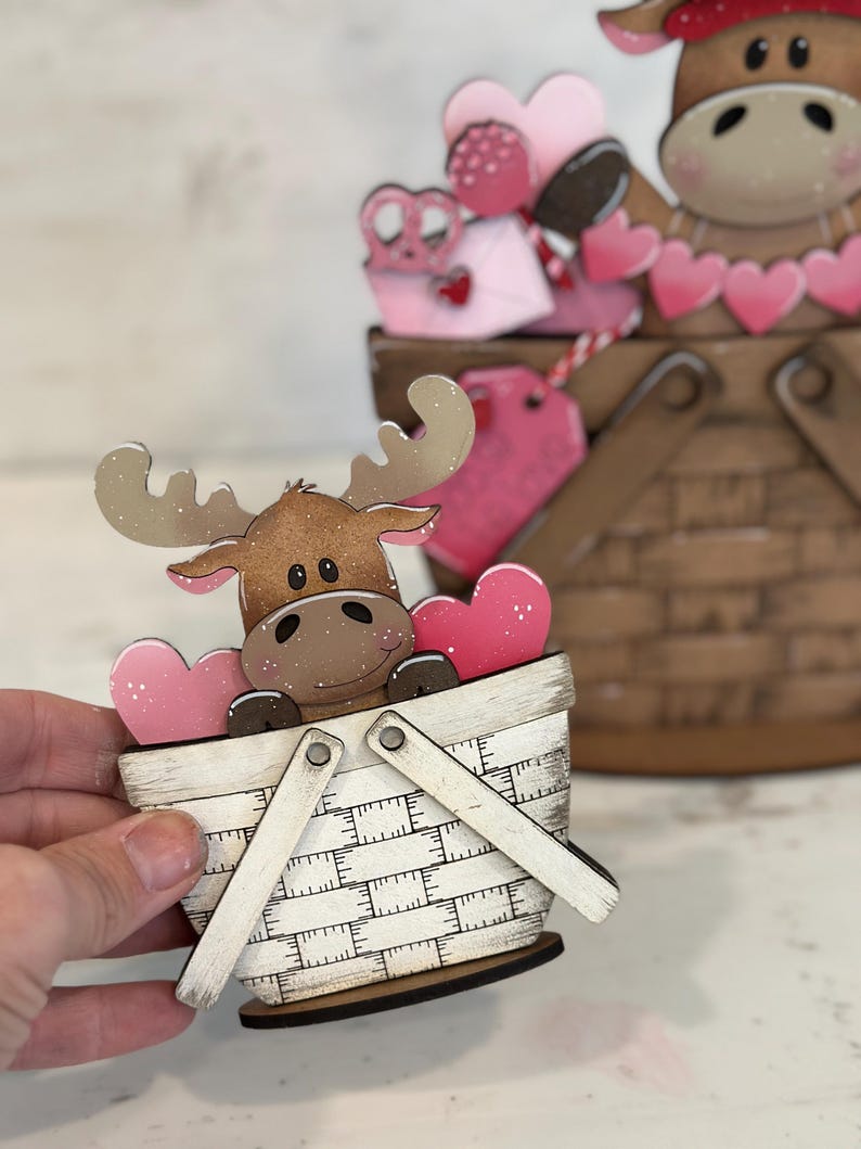 May include: A wooden moose figurine holding pink hearts in a white and brown basket with a white handle. The basket is decorated with a checkered pattern.