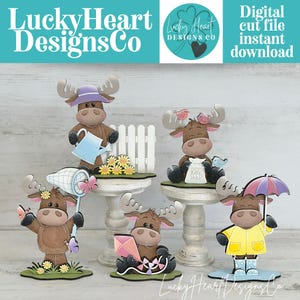 May include: A collection of whimsical moose figurines, each with unique accessories like a watering can, butterfly net, kite, umbrella, and birdseed bag. The figurines are brown with white accents and sit on decorative pedestals. The text "Lucky Heart Designs Co" is visible.