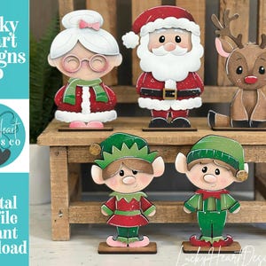 May include: Wooden Christmas figurines including Santa Claus, Mrs. Claus, a reindeer, and two elves. The figurines are painted in festive colors and stand on a small wooden bench. The image also includes the text "Lucky Heart Designs Co" and "Digital cut file instant download".