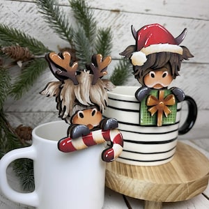 May include: Two decorative wooden mug toppers. One is a Highland cow with antlers and the other is a Highland cow wearing a Santa hat and holding a gift. The first cow is holding a candy cane. Both are perched on white mugs.