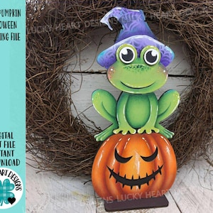 May include: A wooden Halloween decoration featuring a green frog wearing a purple witch hat, perched atop an orange jack-o'-lantern. The frog has large, round eyes and is smiling. The pumpkin has a carved face. Text on the left reads "FROG PUMPKIN HALLOWEEN STANDING FILE".