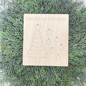 Puede incluir: Un kit de manualidades de madera para hacer un adorno de &aacute;rbol de Navidad. El kit incluye una forma de &aacute;rbol de madera precortada, una estrella y varios adornos. El texto "Build your own tree ornament!" est&aacute; impreso en la madera.