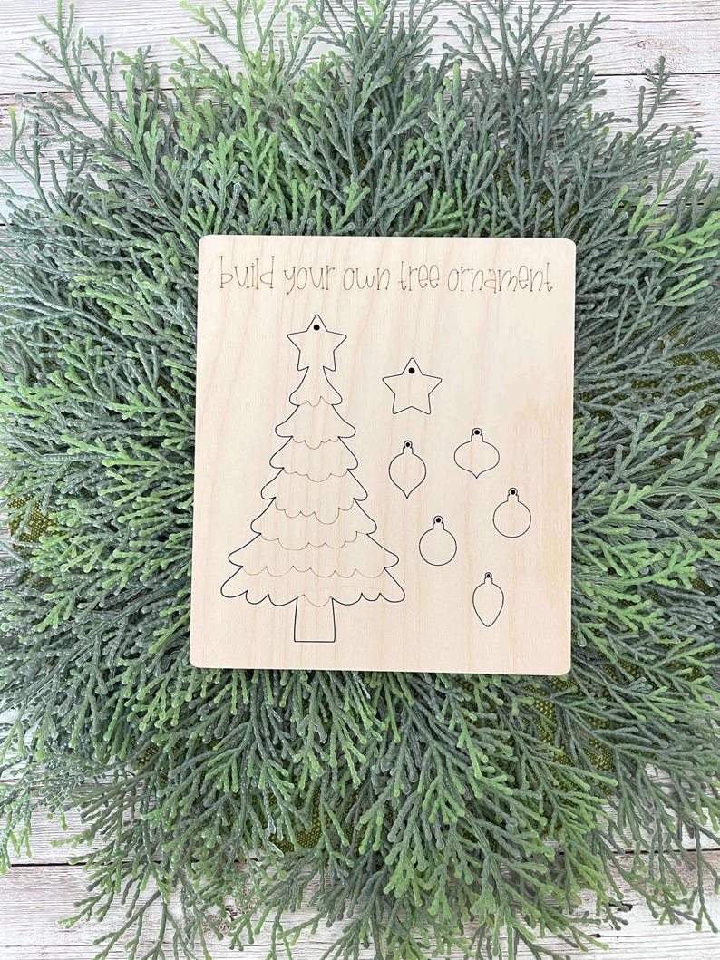 Puede incluir: Un kit de manualidades de madera para hacer un adorno de &aacute;rbol de Navidad. El kit incluye una forma de &aacute;rbol de madera precortada, una estrella y varios adornos. El texto "Build your own tree ornament!" est&aacute; impreso en la madera.