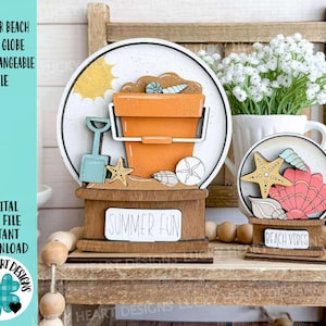 May include: Wooden beach-themed snow globe with interchangeable elements. Features an orange bucket, shovel, starfish, and seashells inside a round frame. The base reads "SUMMER FUN" and "BEACH VIBES". The text "SUMMER BEACH SNOW GLOBE INTERCHANGEABLE FILE" is also visible.