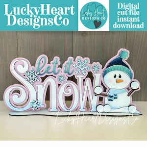 May include: A decorative wooden sign featuring the phrase "Let it Snow" in white and teal, with snowflake accents and a snowman illustration. The sign is in pink and white, with the text "LuckyHeart Designs Co" at the top and bottom.