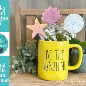 Puede incluir: Una taza amarilla con el texto "BE THE SUNSHINE" contiene recortes decorativos de madera de una estrella de mar, una concha y un dólar de arena. La taza está llena de flores blancas. La imagen también incluye el texto "Lucky Heart Designs Co" y "Digital cut file instant download".