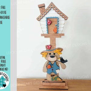 May include: A wooden birdhouse with a bear and scarecrow design. The birdhouse has a small house and chimney. The bear wears a blue vest and a straw hat, holding a black bird. The text "BEAR FALL BIRDHOUSE INTERCHANGEABLE FILE" is on the left.
