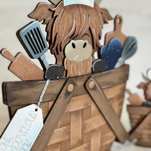 May include: A wooden Highland cow figurine wearing a chef's hat and holding kitchen utensils. The cow is standing in a brown wooden basket with a tag that reads "One Moovelous Cook".