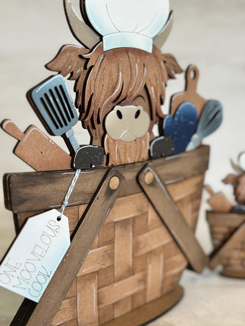 May include: A wooden Highland cow figurine wearing a chef's hat and holding kitchen utensils. The cow is standing in a brown wooden basket with a tag that reads "One Moovelous Cook".