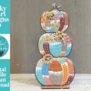 May include: A stacked pumpkin decoration featuring three pumpkins in a vertical arrangement. Each pumpkin is composed of various patterned segments in shades of blue, orange, brown, and pink. Small sunflower embellishments are attached to each.