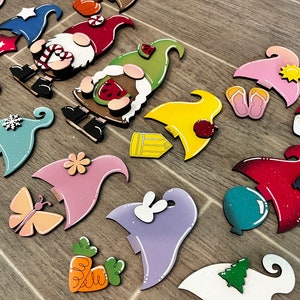 Op de afbeelding: Een verzameling kleurrijke houten dwerg ornamenten met verschillende ontwerpen, waaronder een watermeloen, een vlinder, een wortel, een sneeuwvlok, een zon en een potlood. De ornamenten zijn geschilderd in felle kleuren en hebben een eigenzinnige, handgemaakte uitstraling.