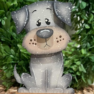 May include: A wooden dog figurine with a gray and white speckled finish. The dog has a large, tan snout and big, round eyes. It is sitting on a small, rectangular base with a brown top and black sides. The background is a green wreath.