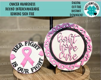 Könnte beinhalten: Zwei runde, rosa und weiße, austauschbare, geneigte Schilder mit einem rosa Band und dem Text "Her Fight Is Our Fight" auf einem Schild und "Fight Hope Cure" auf dem anderen Schild.