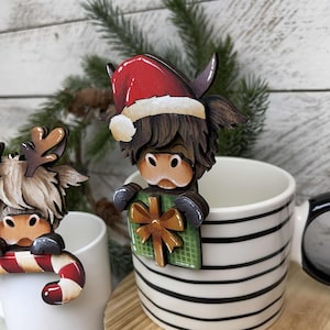 May include: Wooden Christmas ornament of a Highland cow wearing a Santa hat and holding a green gift with a brown bow. The ornament is perched on the rim of a black and white striped mug. A second ornament is visible.