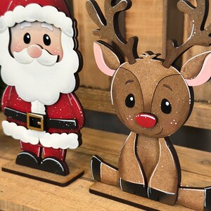 May include: Wooden Christmas decorations featuring Santa Claus and a reindeer. Santa is in a red suit with a white beard and hat. The reindeer has brown fur, antlers, and a red nose. Both are on wooden bases.