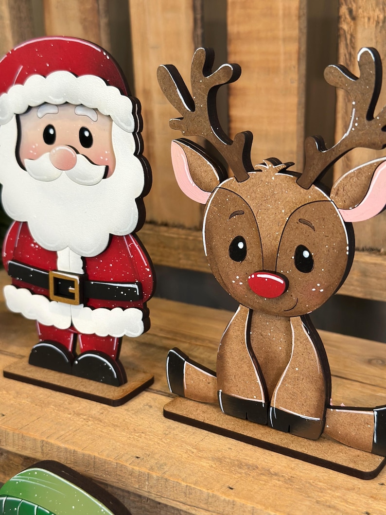 May include: Wooden Christmas decorations featuring Santa Claus and a reindeer. Santa is in a red suit with a white beard and hat. The reindeer has brown fur, antlers, and a red nose. Both are on wooden bases.