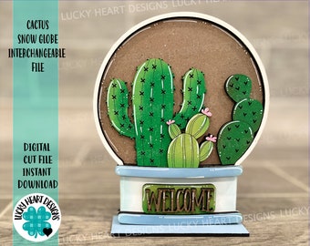 May include: A wooden cactus snow globe with a welcome sign. The snow globe features a brown base with a light blue top and a clear dome. The cactus is green with pink flowers and is surrounded by smaller cactus plants.