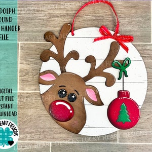 May include: A round wooden door hanger featuring a cartoon Rudolph the Red-Nosed Reindeer with a red nose and a green Christmas bauble hanging from his antlers. The background is a white woodgrain pattern.