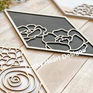 May include: Wooden wall art pieces with floral designs. One piece features a black background with white floral outlines, while others have natural wood tones. The pieces are diamond-shaped and have a layered effect.