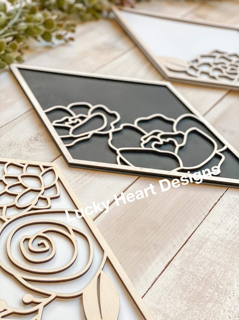 May include: Wooden wall art pieces with floral designs. One piece features a black background with white floral outlines, while others have natural wood tones. The pieces are diamond-shaped and have a layered effect.