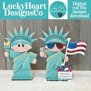 May include: Two wooden Statue of Liberty figures. One holds a torch and a book with "JULY IV" on it. The other wears star-spangled sunglasses and holds a flag. A small crate holds fireworks. The text "Lucky Heart Designs Co" is at the top and bottom.