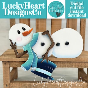 以下が含まれることがあります： 青いスカーフ、ニンジンの鼻、黒い目の木製雪だるまの装飾。 雪だるまは青いシルクハットをかぶっています。 「LuckyHeart Designs Co」の文字が見えます。