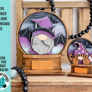 May include: Wooden Halloween snow globe decorations. The larger globe features a gnome with a purple hat and bat wings, with the word "SPOOKTACULAR" on the base. The smaller globe has a gnome with a hat and the word "BOO".