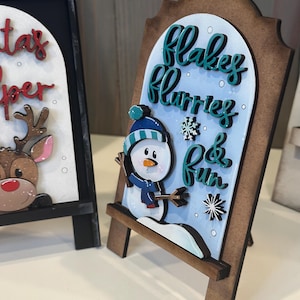 May include: A wooden sign with a snowman graphic and the words "Flakes, Flurries & Fun" in teal script. The sign has a brown frame and a light blue background. A second sign with a reindeer is partially visible.
