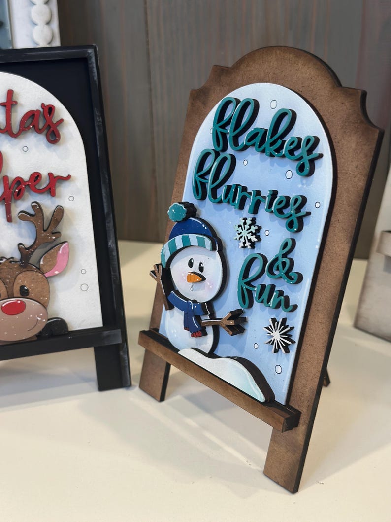 May include: A wooden sign with a snowman graphic and the words "Flakes, Flurries & Fun" in teal script. The sign has a brown frame and a light blue background. A second sign with a reindeer is partially visible.