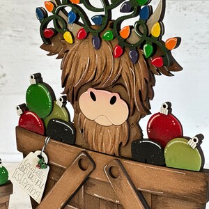 K&ouml;nnte beinhalten: Ein h&ouml;lzerner Weihnachtsanh&auml;nger mit einer schottischen Hochlandkuh, die einen Kranz aus bunten Lichtern tr&auml;gt und einen Korb voller Kugeln tr&auml;gt. Der Anh&auml;nger hat ein Schild mit der Aufschrift "Cowbells are you listening".