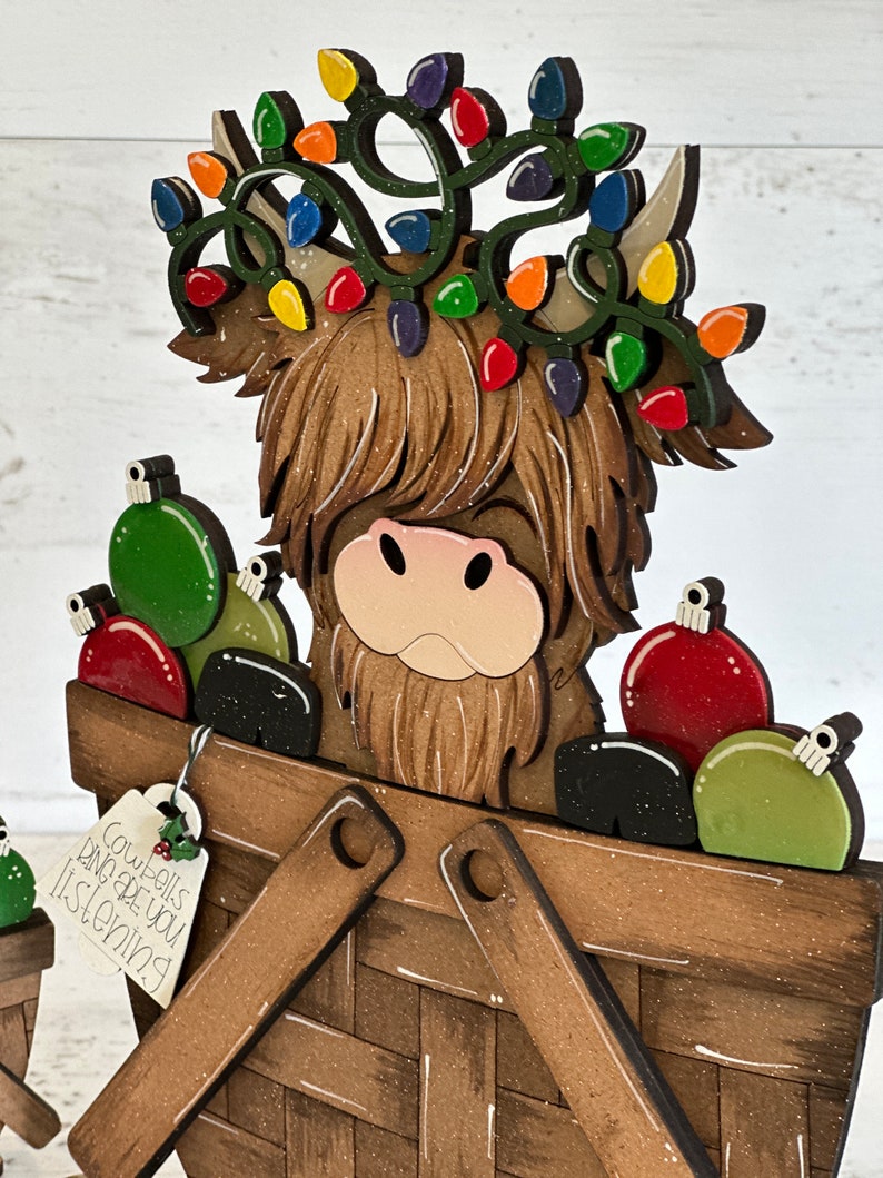 K&ouml;nnte beinhalten: Ein h&ouml;lzerner Weihnachtsanh&auml;nger mit einer schottischen Hochlandkuh, die einen Kranz aus bunten Lichtern tr&auml;gt und einen Korb voller Kugeln tr&auml;gt. Der Anh&auml;nger hat ein Schild mit der Aufschrift "Cowbells are you listening".