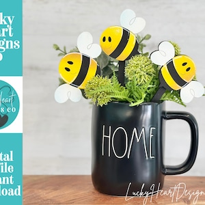 May include: A black mug with the word "HOME" in white lettering holds greenery and three wooden bee decorations. The bees are yellow and black with white wings. The image also includes the text "Lucky Heart Designs Co" and "Digital cut file instant download."