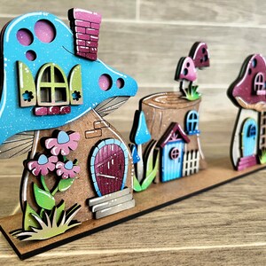 以下が含まれることがあります： 3つのキノコの家を持つ木製の妖精の家セット。最大のキノコの家は、ピンクと白のドアと緑の屋根が付いた青いキノコです。真ん中の家は、小さな青いドアと赤い屋根が付いた木の切り株です。最小の家は、青いドアが付いた赤いキノコです。家はすべて木製の土台の上にあります。