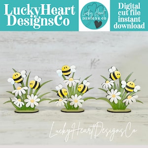Puede incluir: Recortes de madera de abejas y margaritas. Cada arreglo presenta abejas amarillas y negras que flotan sobre margaritas blancas con centros amarillos y hojas verdes. El texto "Lucky Heart Designs Co" es visible.