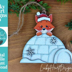 May include: A wooden Christmas ornament featuring a cartoon fox wearing a Santa hat, peeking over a light blue igloo. The ornament has snowflake details and is suspended by a twine loop. The text "Lucky Heart Designs Co" is visible.