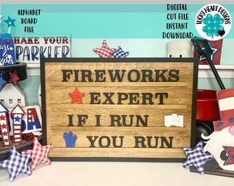 May include: A wooden sign with the text "FIREWORKS EXPERT IF I RUN YOU RUN" in black letters. The sign is decorated with red, white, and blue stars and a blue firework graphic. The sign is surrounded by red, white, and blue decorations.