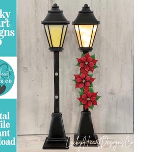 May include: Two black decorative lampposts with illuminated yellow lanterns. One lamppost is decorated with three red poinsettia flowers and green leaves. The text "Lucky Heart Designs Co" and "Digital cut file instant download" are visible.
