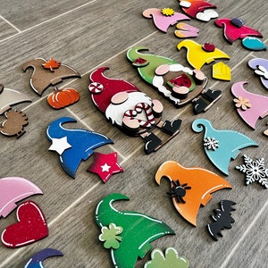 Op de afbeelding: Een verzameling van 18 onafgewerkte houten dwerg ornamenten met verschillende beschilderde ontwerpen. De dwergen zijn in verschillende kleuren geschilderd, waaronder rood, blauw, groen, roze en oranje. Elke dwerg heeft een ander ontwerp, zoals een pompoen, een ster, een hart, een sneeuwvlok, een spin, een vlinder, een klavertjevier en een snoepstok.