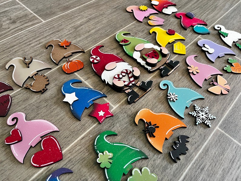 Op de afbeelding: Een verzameling van 18 onafgewerkte houten dwerg ornamenten met verschillende beschilderde ontwerpen. De dwergen zijn in verschillende kleuren geschilderd, waaronder rood, blauw, groen, roze en oranje. Elke dwerg heeft een ander ontwerp, zoals een pompoen, een ster, een hart, een sneeuwvlok, een spin, een vlinder, een klavertjevier en een snoepstok.