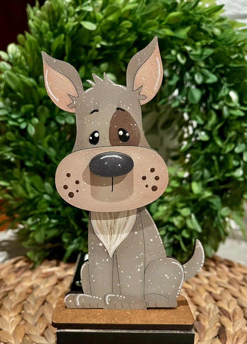 May include: A wooden dog figurine with a brown and gray color scheme. The dog has a large snout, floppy ears, and a brown spot over one eye. The figurine is sitting on a small wooden base, with a green leafy background.