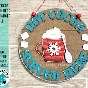 Puede incluir: Un colgador de puerta redondo de madera con el texto "Hot Cocoa Served Here" en letras azul turquesa. El colgador presenta una taza roja con un diseño de copo de nieve, crema batida blanca y una cuchara blanca. También hay dos malvaviscos blancos a cada lado de la taza.