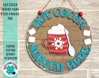 May include: A round wooden door hanger with the text "Hot Cocoa Served Here" in teal blue letters. The hanger features a red mug with a snowflake design, white whipped cream, and a white spoon. There are also two white marshmallows on either side of the mug.