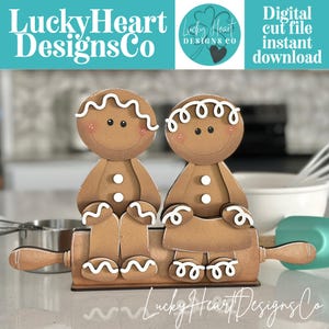 May include: A wooden gingerbread couple figurine sitting on a rolling pin base. The figures are light brown with white icing details on their heads, hands, and feet, and two white buttons on their chests. The text LuckyHeartDesignsCo is visible.
